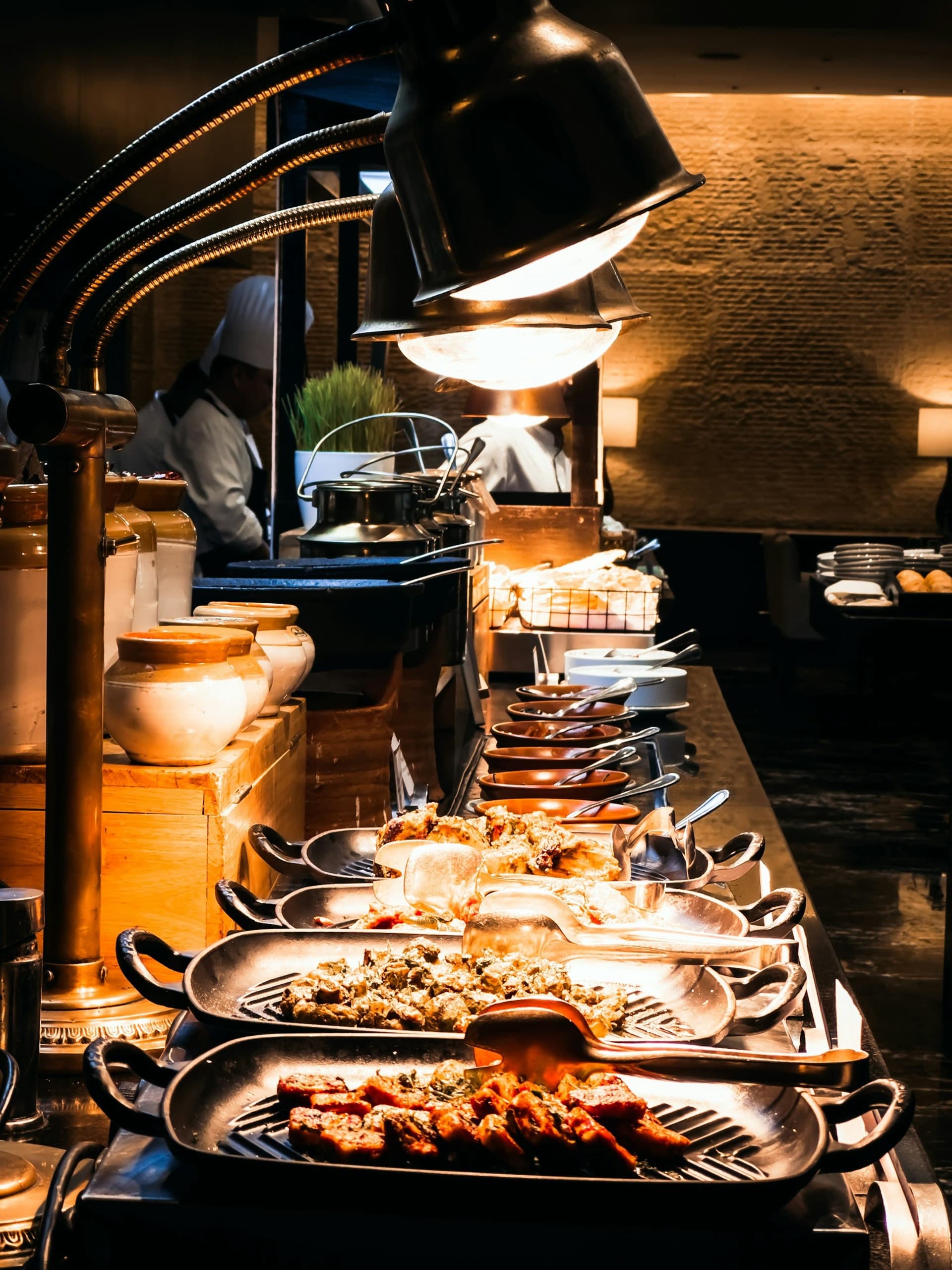 Ein Buffet mit warmen Speisen in beheizten Behältern unter Wärmelampen, im Hintergrund ein Koch.