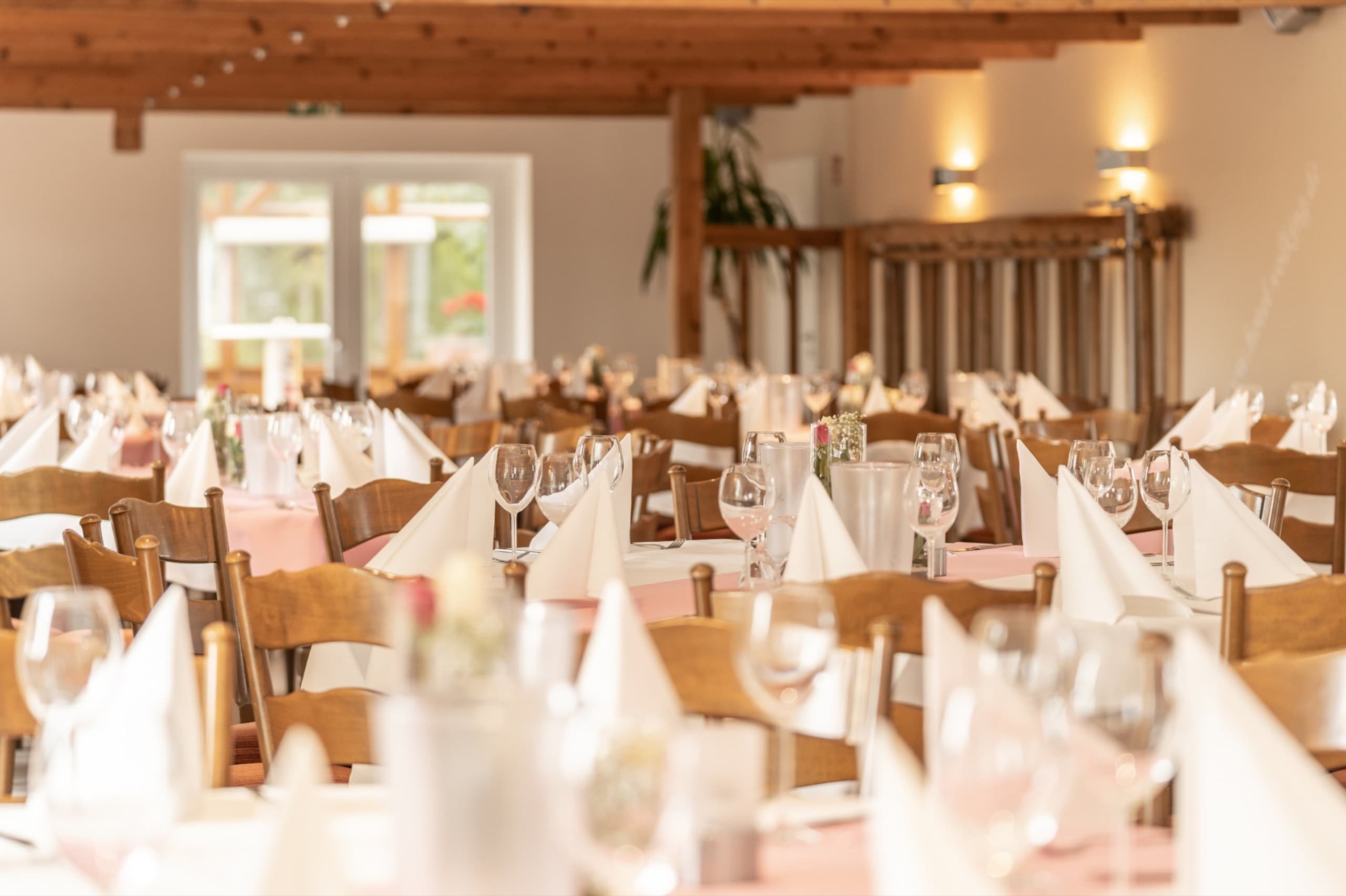 Festlich gedeckter Saal mit Holztischen, Stühlen, Gläsern und weißen Servietten, bereit für eine Feier.