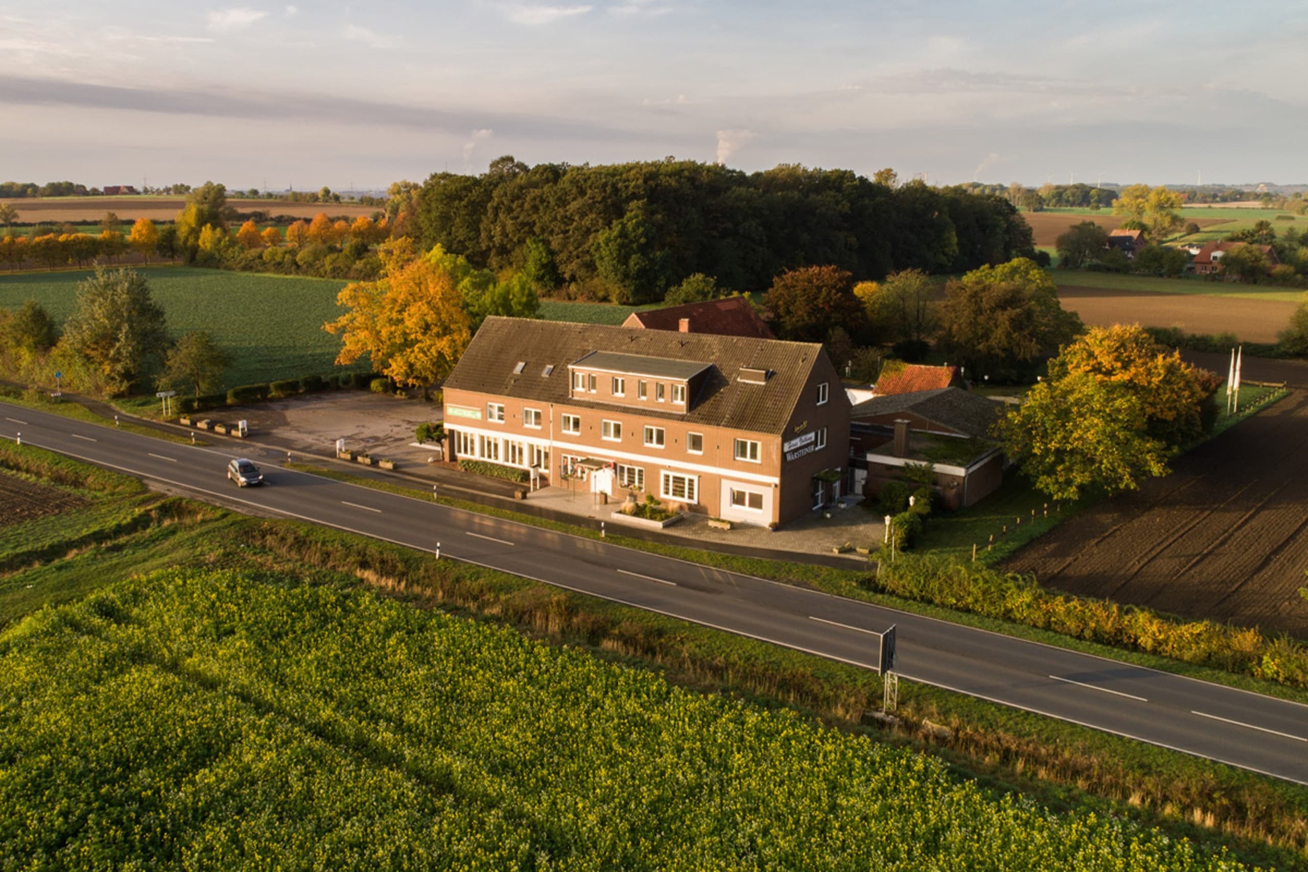 Ein Hotelgebäude mit roten Ziegeln und grauem Dach liegt neben einer Straße, umgeben von grünen Feldern und Bäumen unter blauem Himmel.